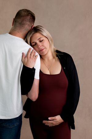 Pregnant woman in burgundy dress leans on partner's shoulder, cradling her baby bump during a maternity photo session.