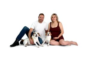Couple sitting on white background with two beagle dogs, woman in burgundy dress appears pregnant, man in white shirt and jeans.