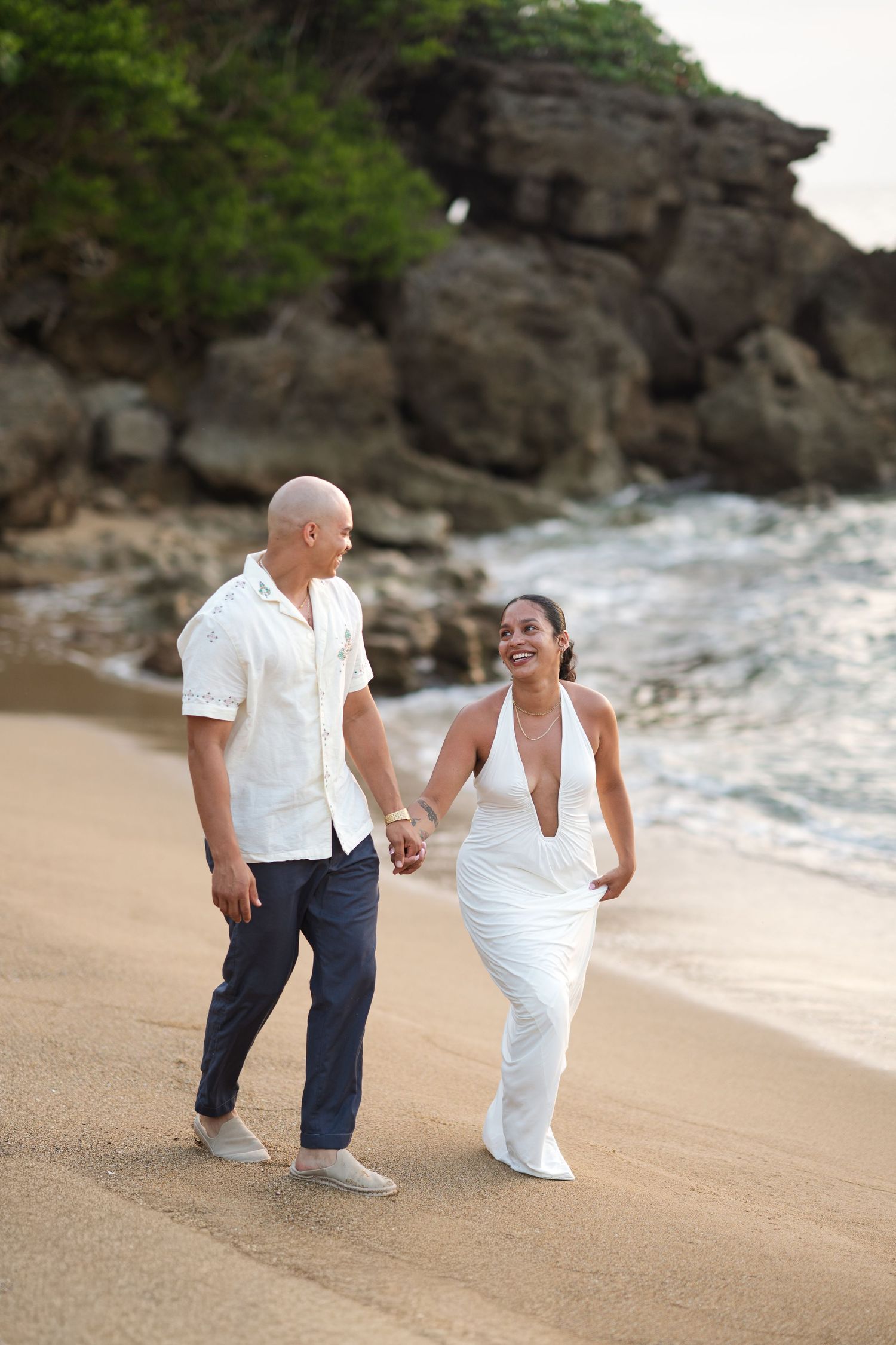 Couple holding hands walking on sandy beach, man in white shirt and navy pants, woman in white halter dress, rocky cliffs behind.