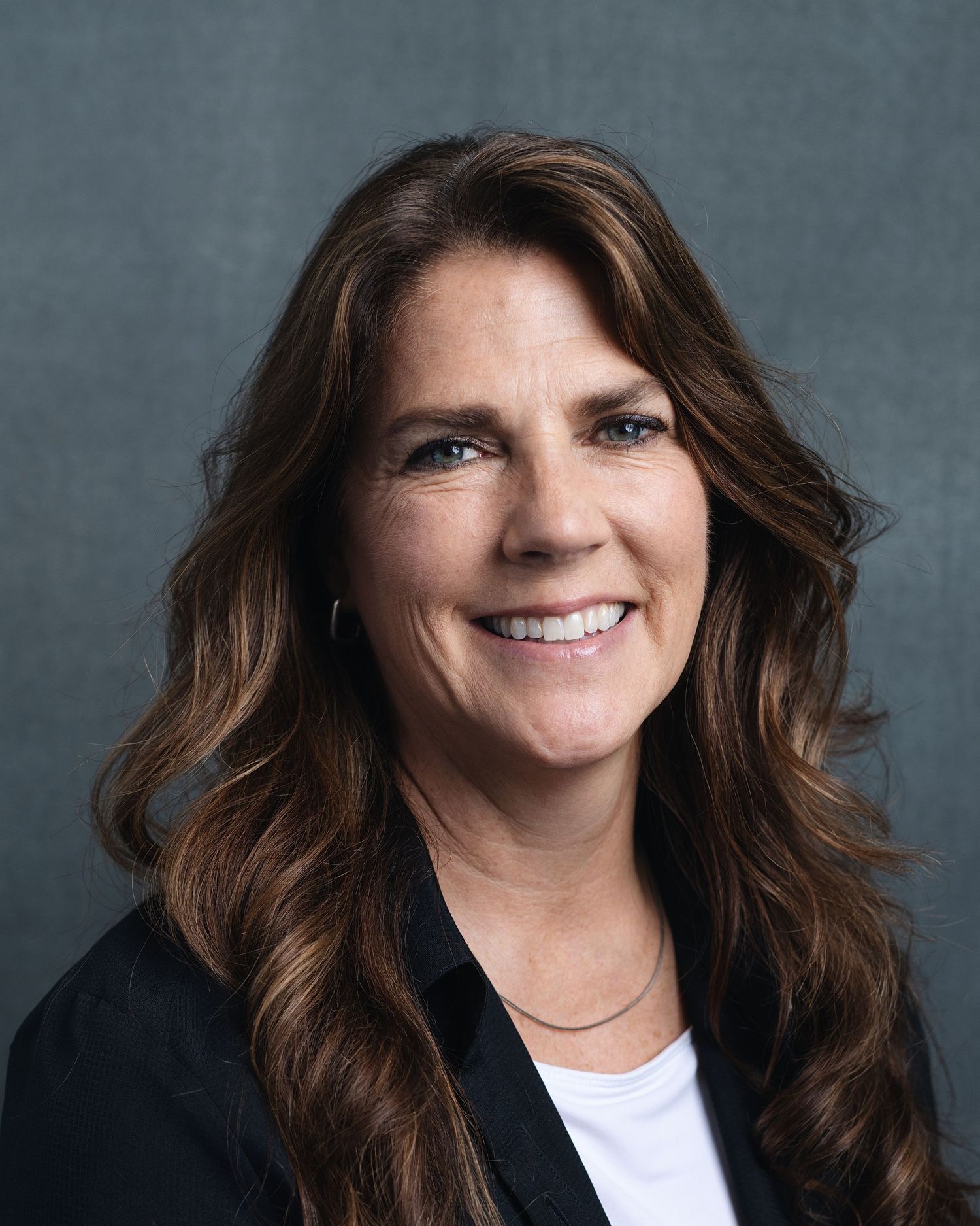 Professional headshot of a smiling woman with long brown wavy hair wearing a black blazer against a gray background.