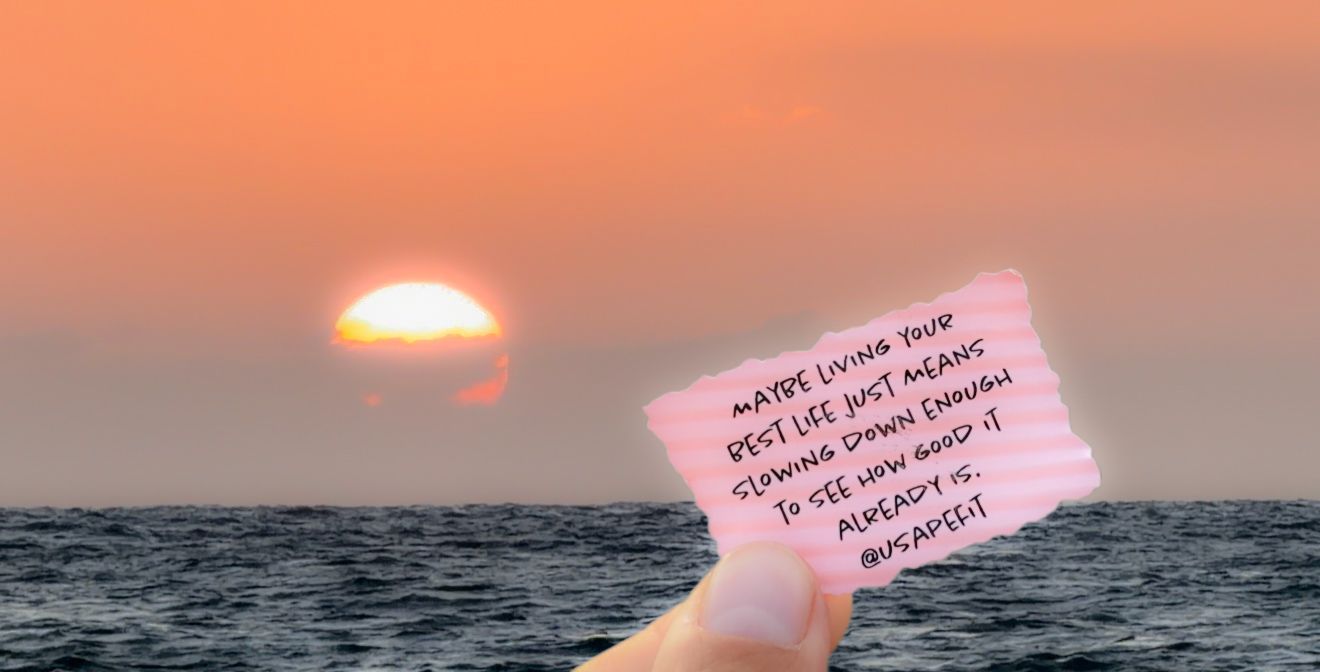 Hand holding a note card at sunset over the ocean with a pink sky and glowing sun on the horizon.