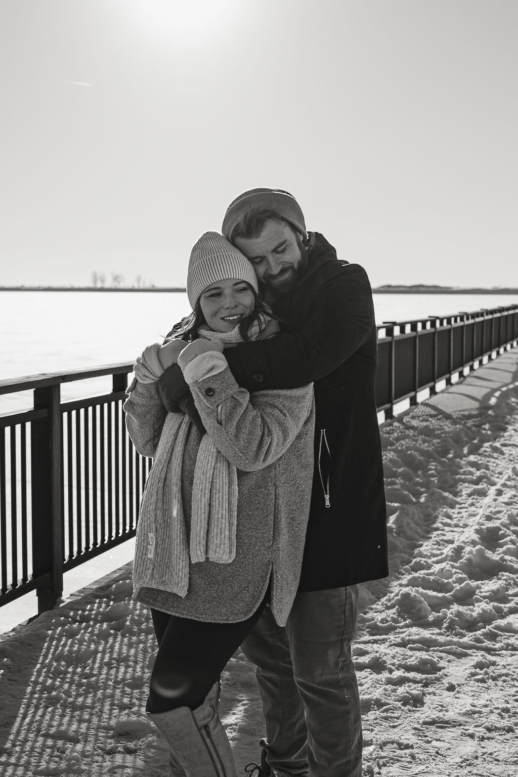 Black and white photo of a couple embracing on a snowy waterfront promenade in winter coats and hats.