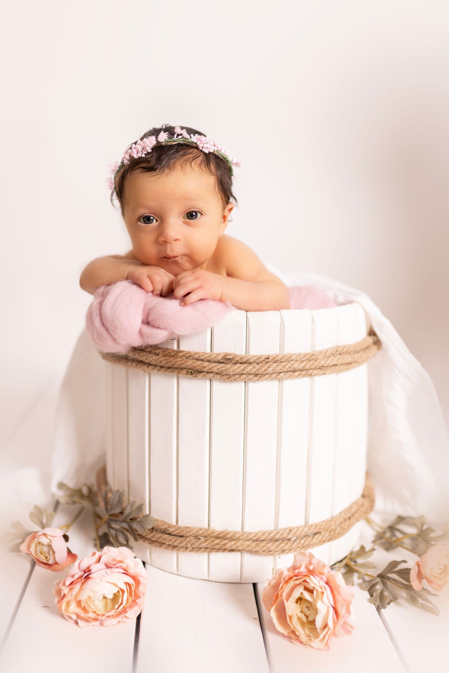 Baby wearing pink headband sits in white wooden barrel prop with peach roses for newborn photography session.