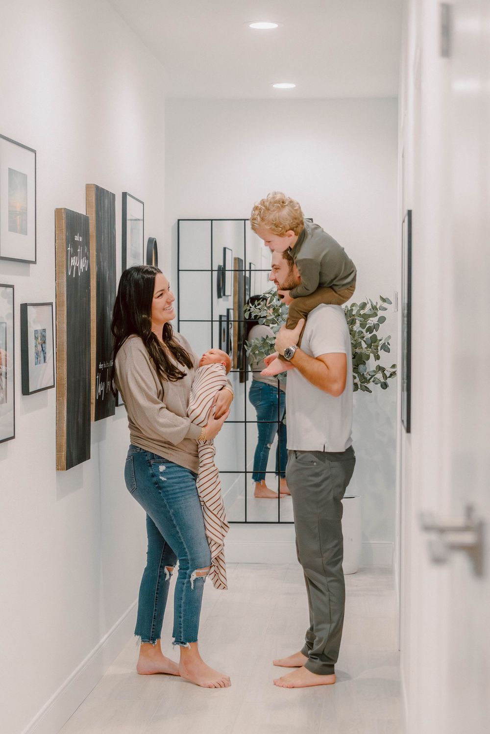 A happy family in a modern hallway, mom holding a newborn, dad with toddler on shoulders, framed art on white walls.