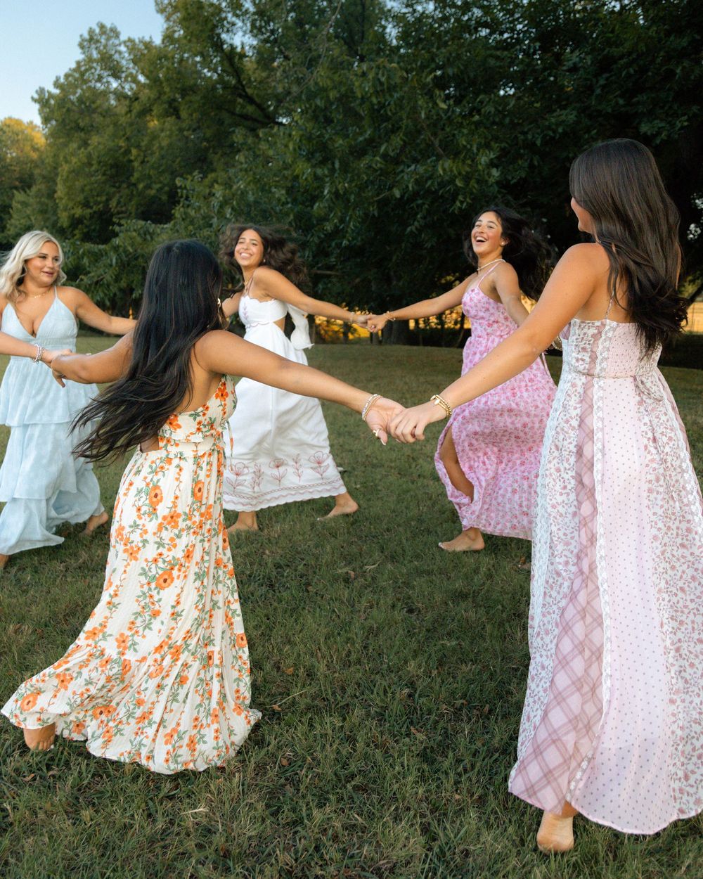 Five women in floral maxi dresses hold hands and dance in a circle outdoors on a grassy field.