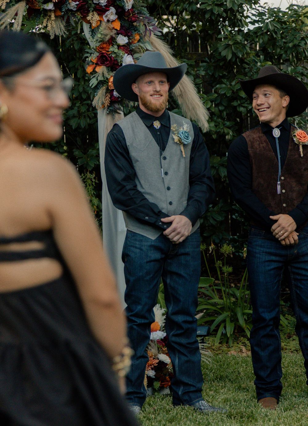 Groom in cowboy hat and gray vest smiles at bride during outdoor western-themed wedding ceremony.