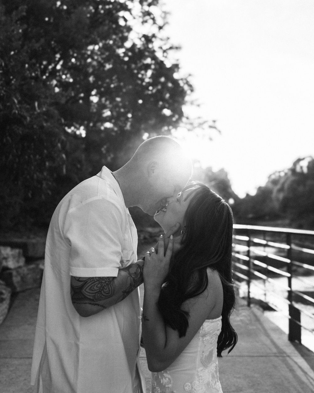 Black and white photo of a couple sharing a romantic moment on a bridge, backlit by golden sunlight.