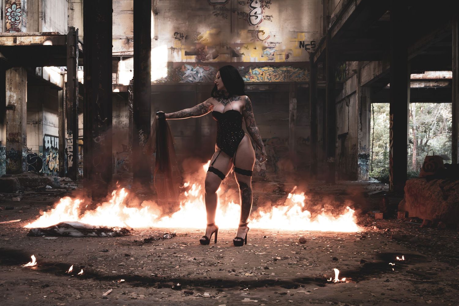 A woman in black outfit poses dramatically among flames in an abandoned graffiti-covered industrial building at night.