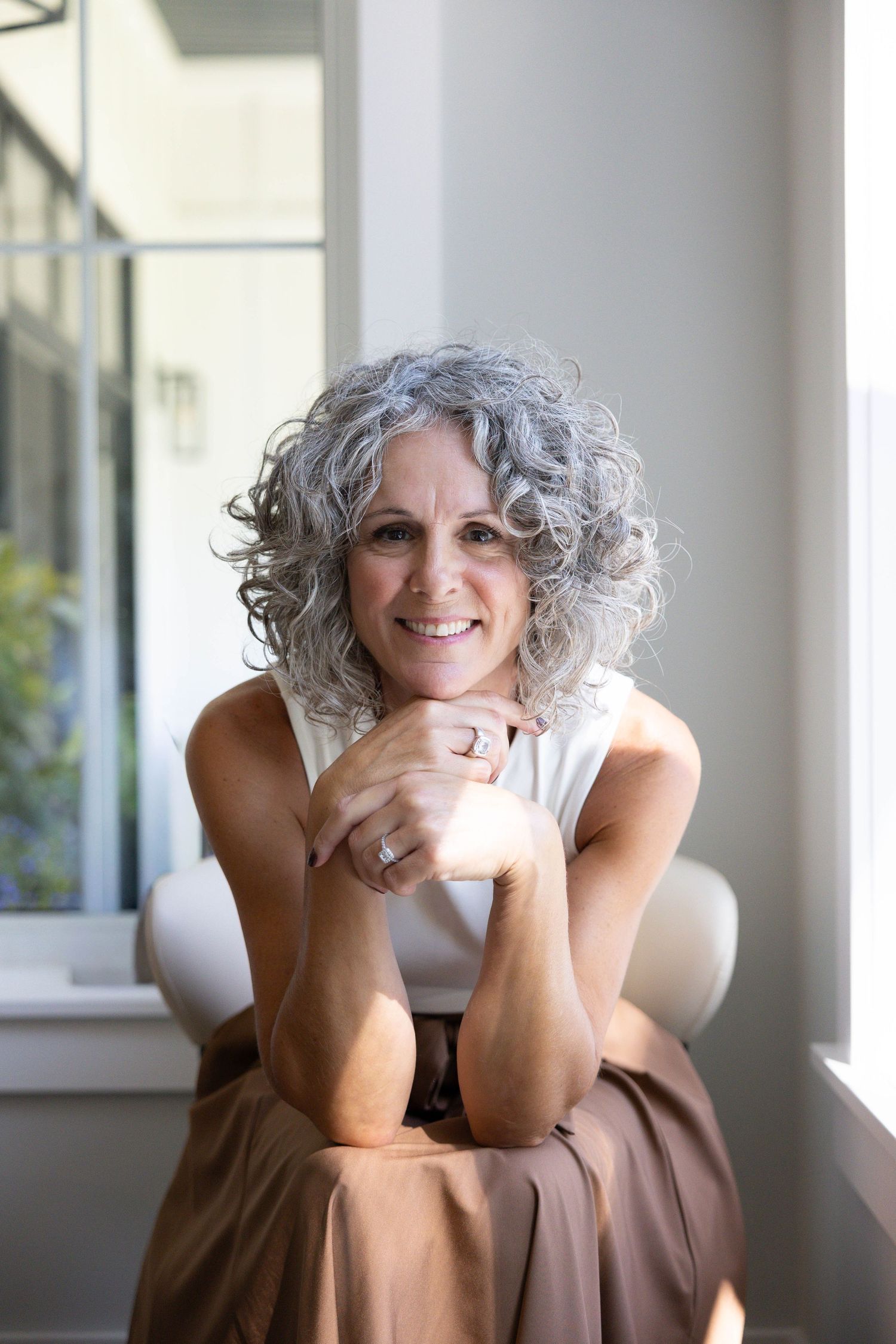Smiling middle-aged woman with curly gray hair sitting relaxed near a bright window, hands clasped, wearing a sleeveless top.