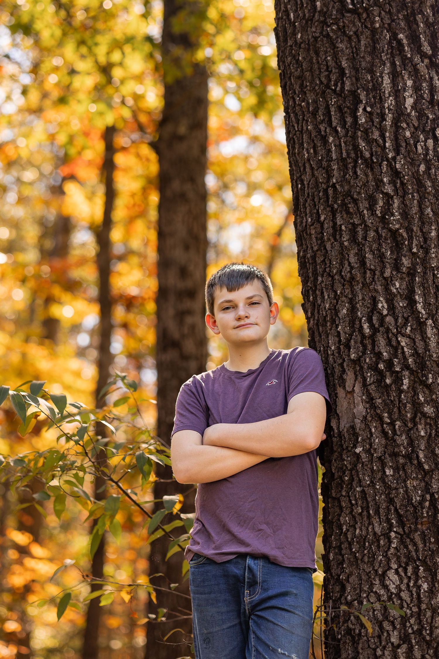 A teenage boy in a purple shirt and jeans leans against a tree in an autumn forest with golden foliage.