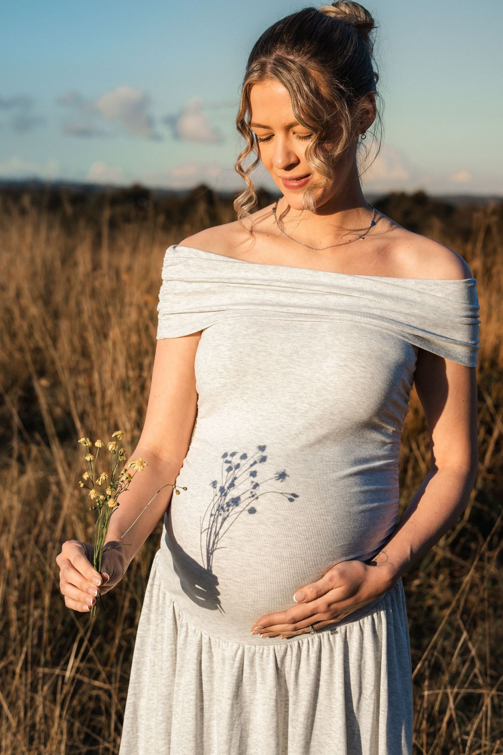 Pregnant woman in gray off-shoulder dress holding wildflowers in golden field, cradling baby bump with floral belly art.