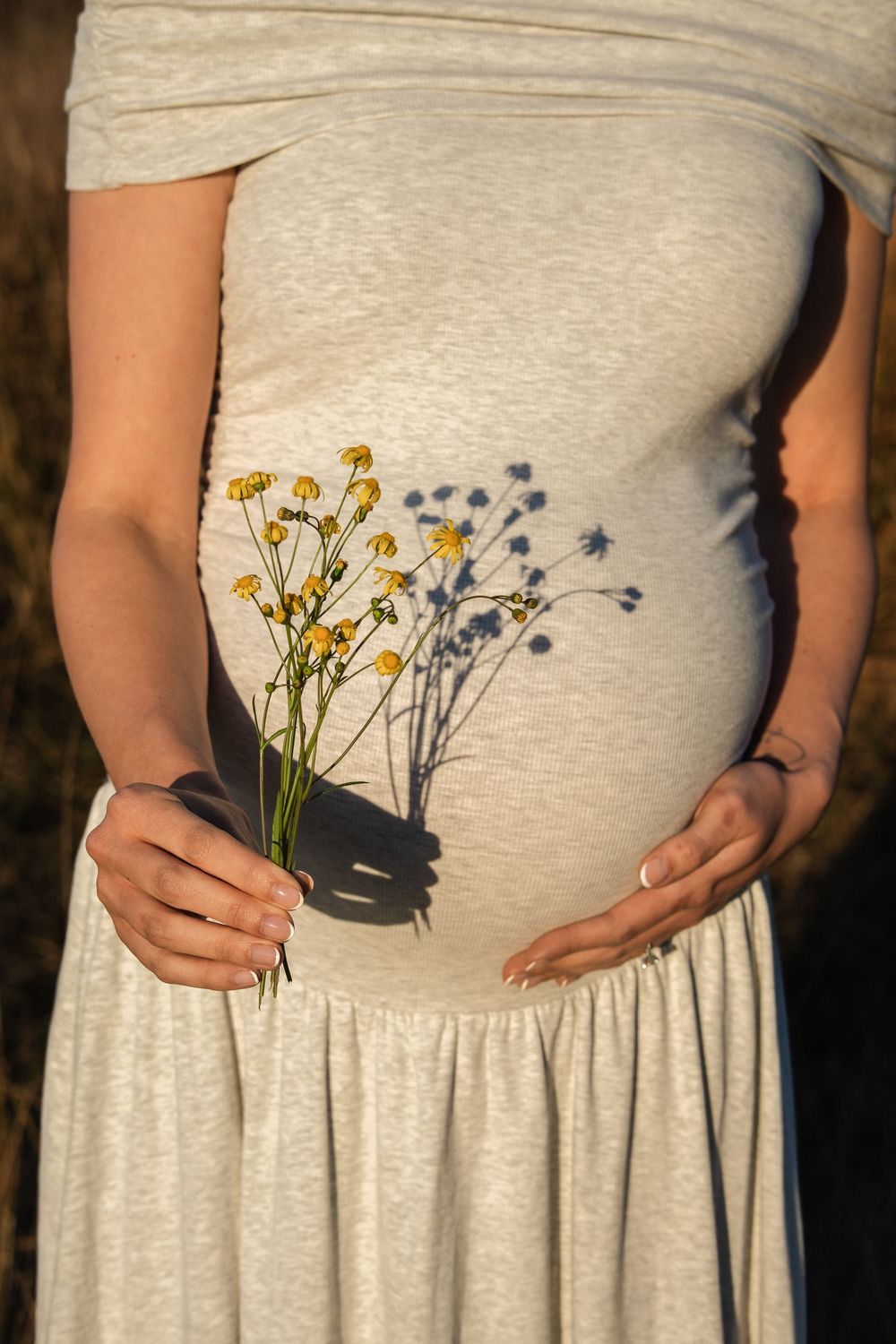 Pregnant woman in gray dress holding yellow wildflowers against her baby bump in warm golden sunlight.