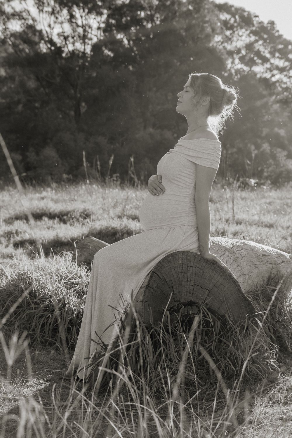Pregnant woman in elegant dress sits on log in field, black and white maternity photo with soft natural light.