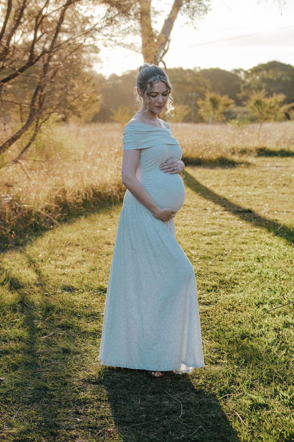 Pregnant woman in white off-shoulder gown cradling baby bump in golden sunset field.
