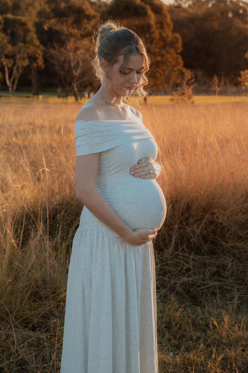 Pregnant woman in white off-shoulder gown cradling baby bump at golden sunset in grassy field at Rouse Hill Regional Park.