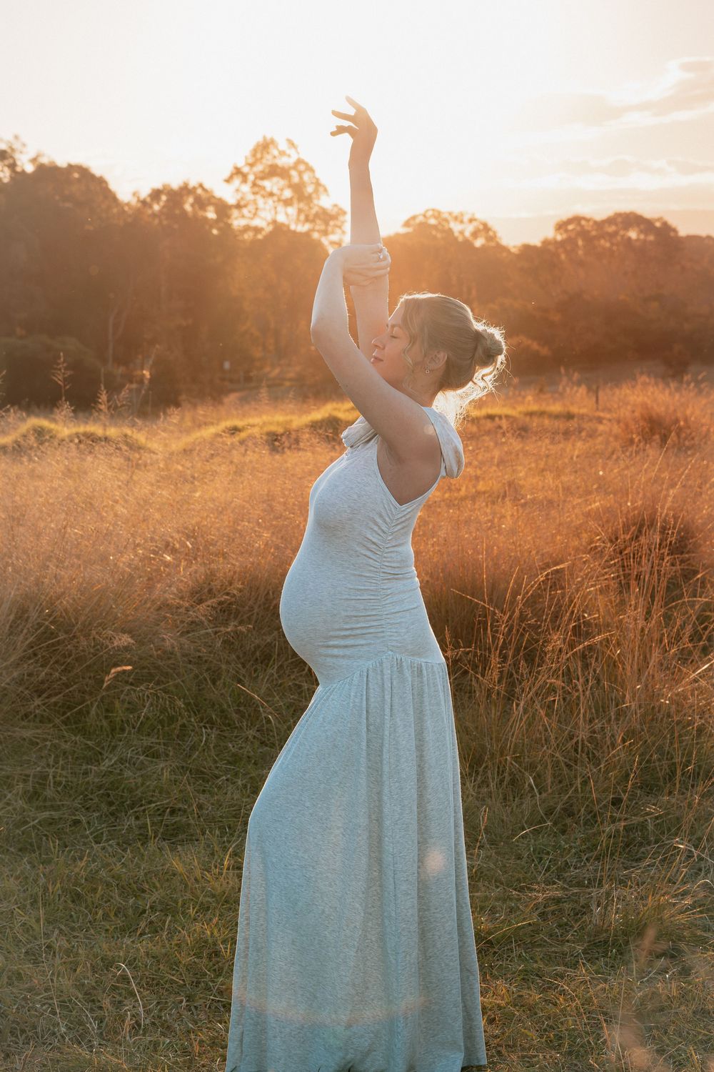 Pregnant woman in white dress posing gracefully at golden sunset in a field.