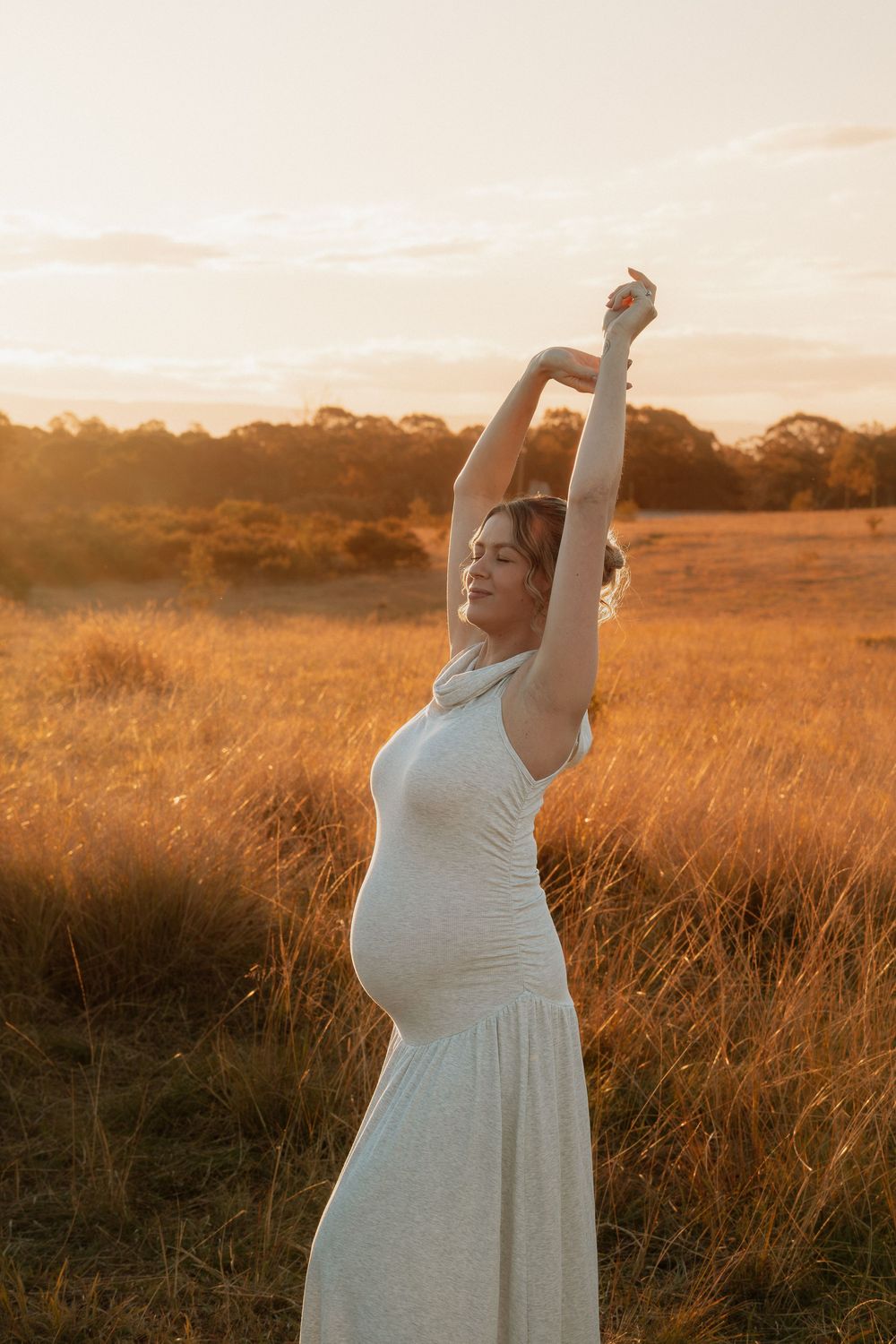 Pregnant woman in white dress stands in golden field at sunset, arms raised joyfully, glowing in warm light.