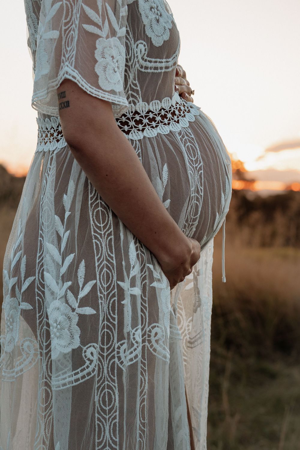 Pregnant woman in sheer white lace dress holding her baby bump at golden hour in a field during maternity photoshoot in Rouse Hill.