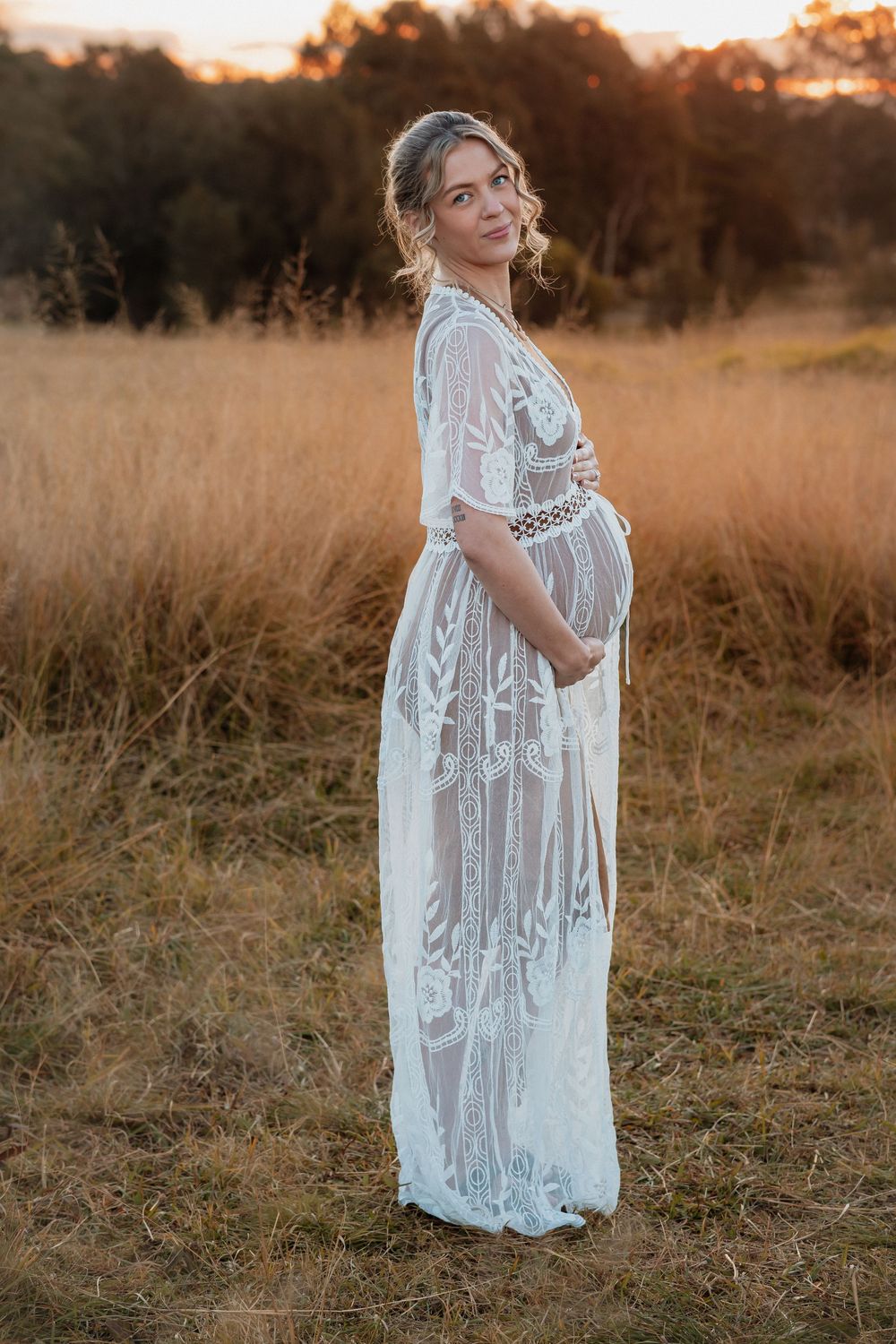 Pregnant woman in white lace boho maxi dress standing in golden field at sunset, cradling baby bump.