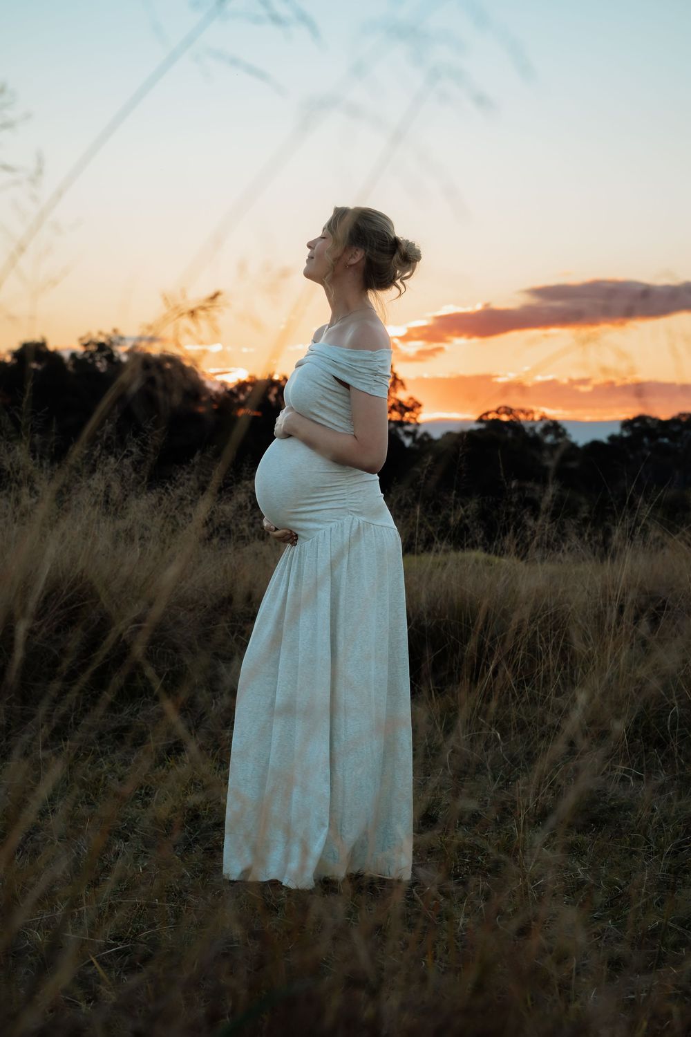 Pregnant woman in white off-shoulder dress standing in golden grass field at sunset, cradling her baby bump.