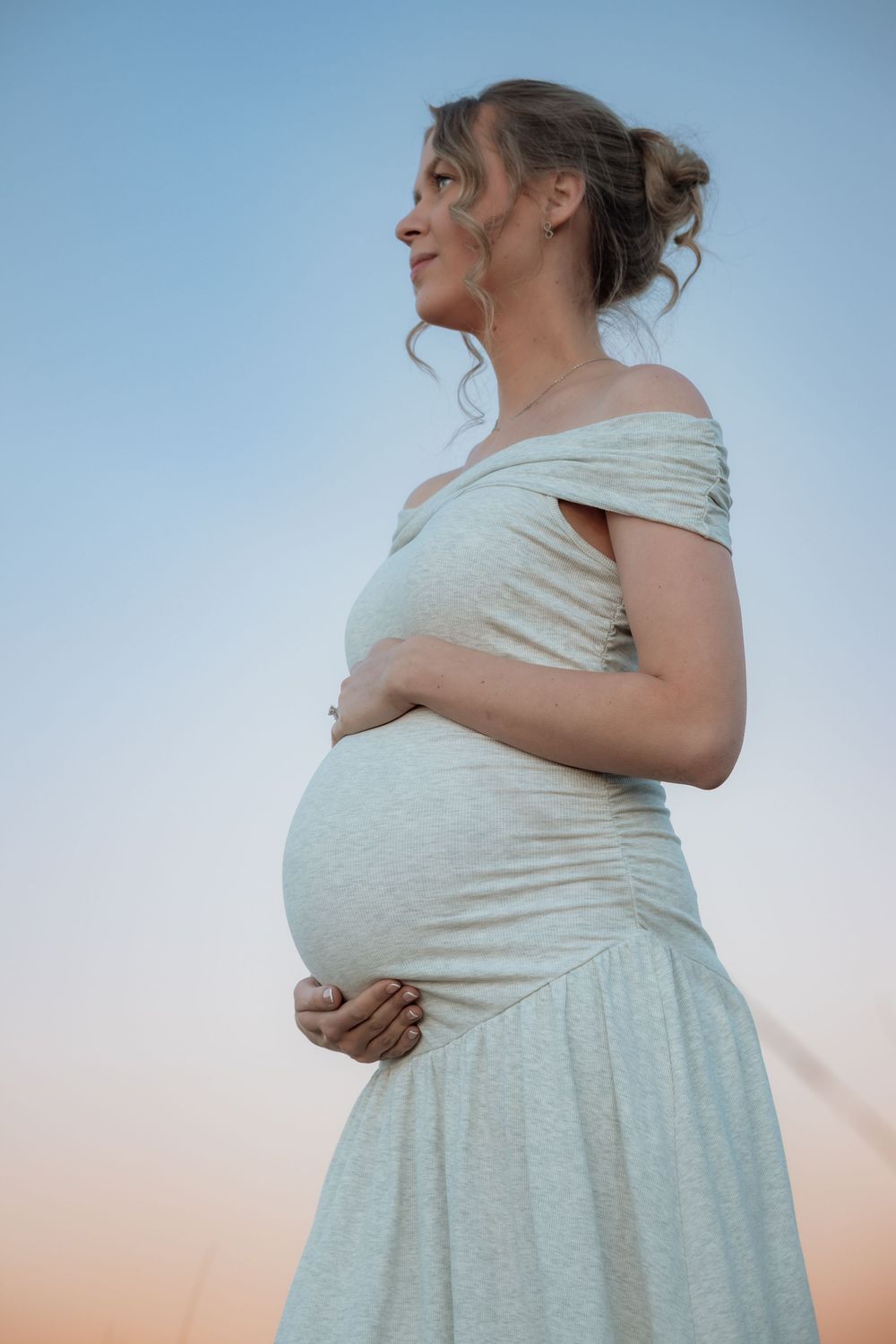 Pregnant woman in white off-shoulder dress cradling her baby bump against a soft blue sky at sunset.
