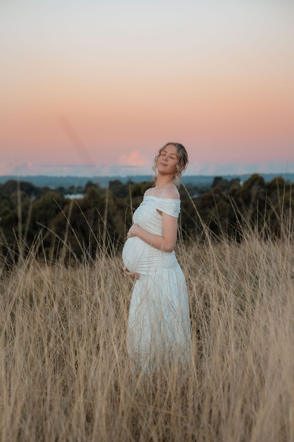 Pregnant woman in white off-shoulder dress standing in golden grass field at sunset, cradling baby bump.
