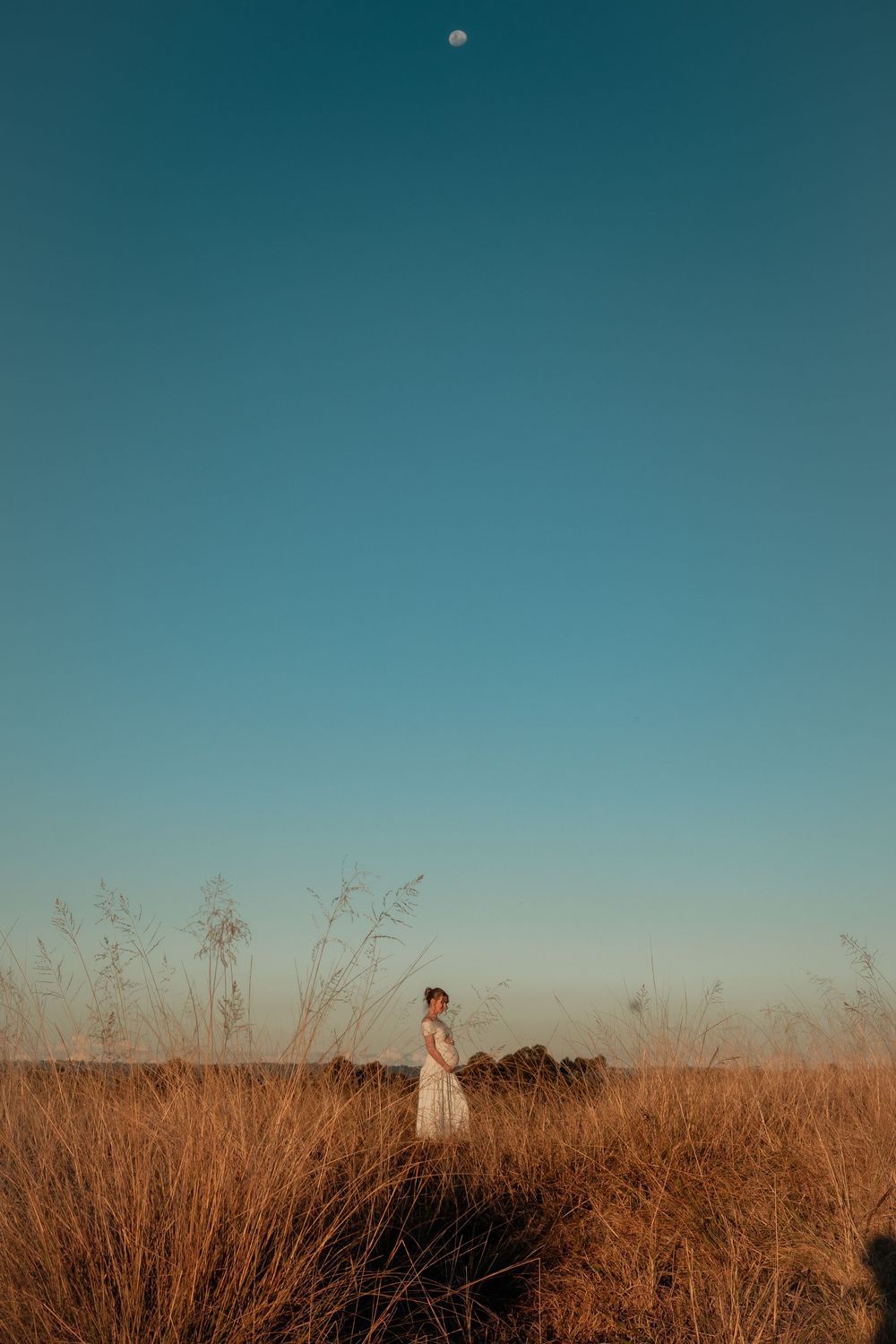 Woman in white dress standing in golden grass field under clear blue sky with moon visible at dusk.