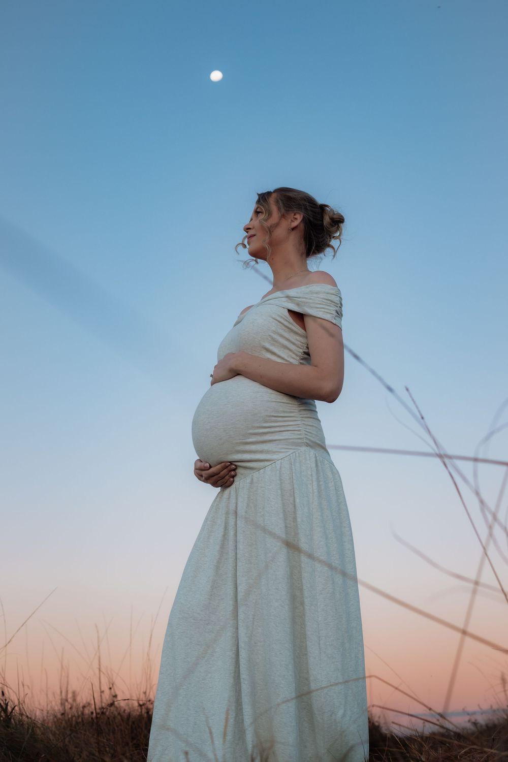Pregnant woman in white off-shoulder dress cradling belly at dusk with moon in blue-pink sky In Rouse Hill.