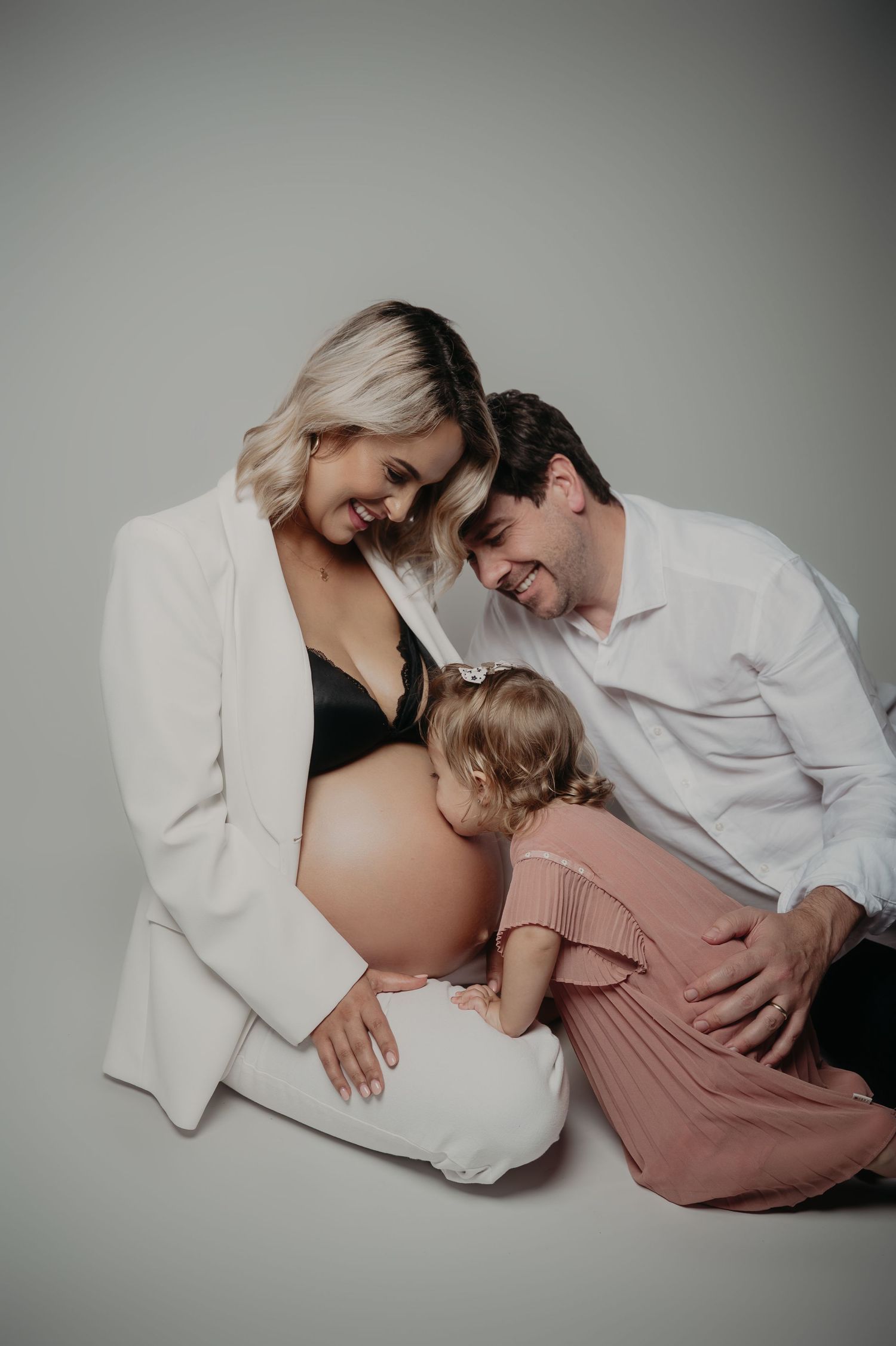 Pregnant woman in white blazer sits with husband and toddler daughter touching her baby bump in studio maternity photo.