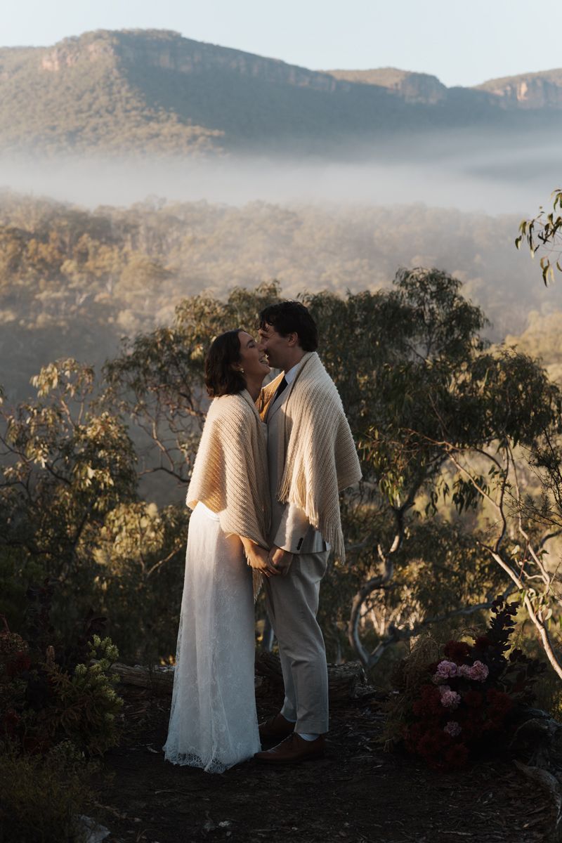 A couple shares a kiss at a misty blue mountain overlook, bride in white dress with shawl, groom in gray suit elopement photoshoot