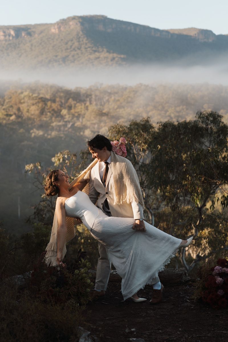 A couple shares a romantic dip dance in wedding attire with misty mountains and eucalyptus trees in the background. Elopement photoshoot
