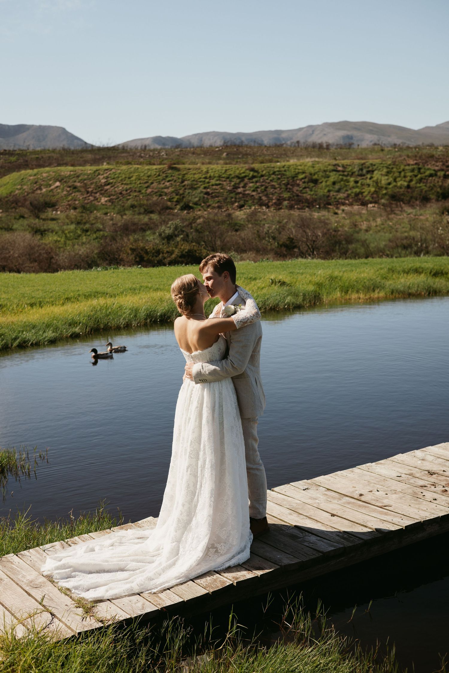 Bride and groom embracing on wooden dock beside serene lake with mountain backdrop.