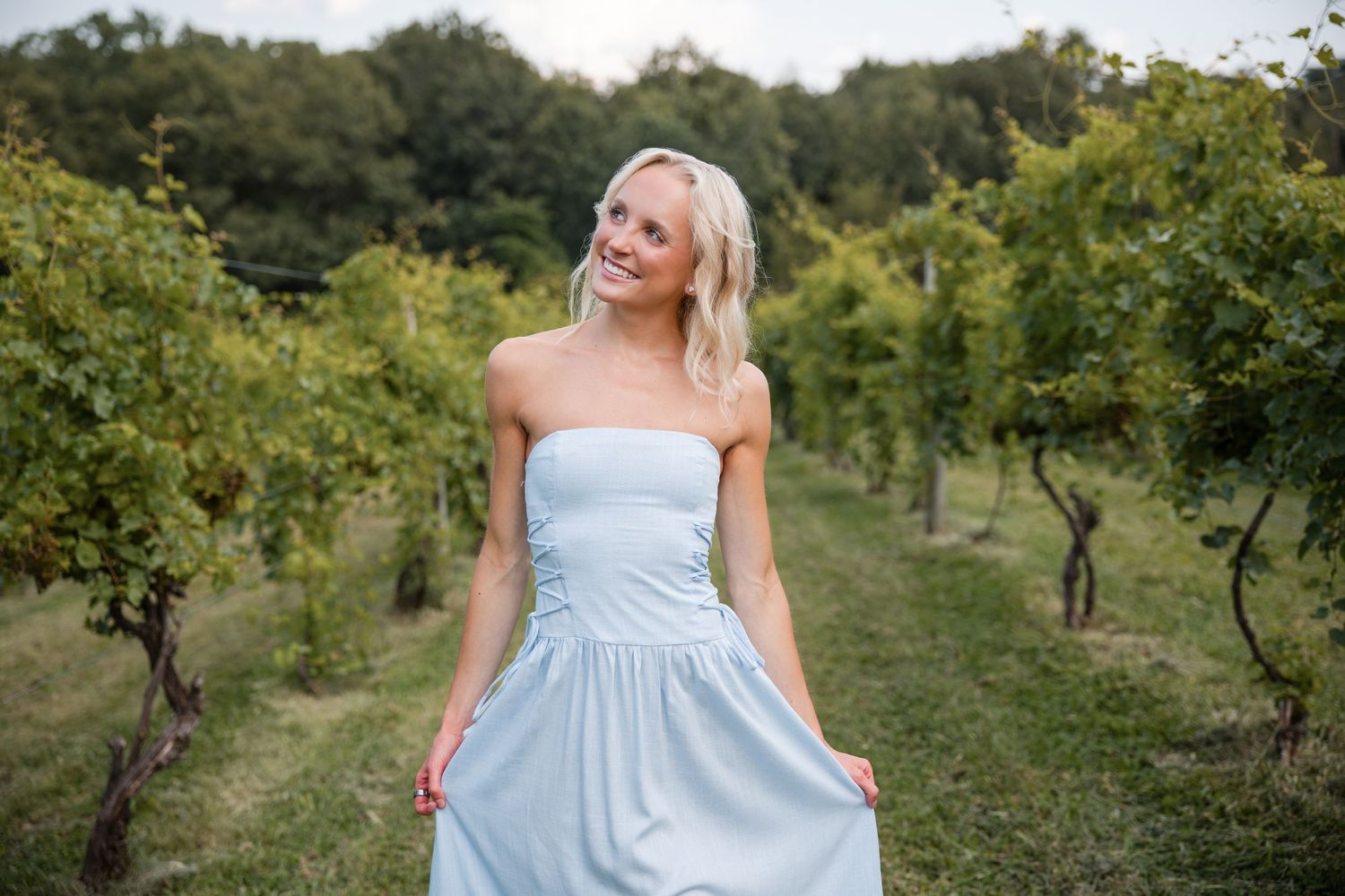 Smiling blonde woman in a light blue strapless dress standing among vineyard rows on a cloudy day.