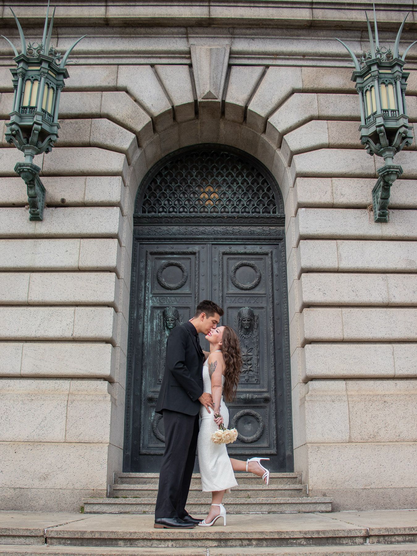 A couple shares a kiss in front of an ornate arched door at a grand stone building.