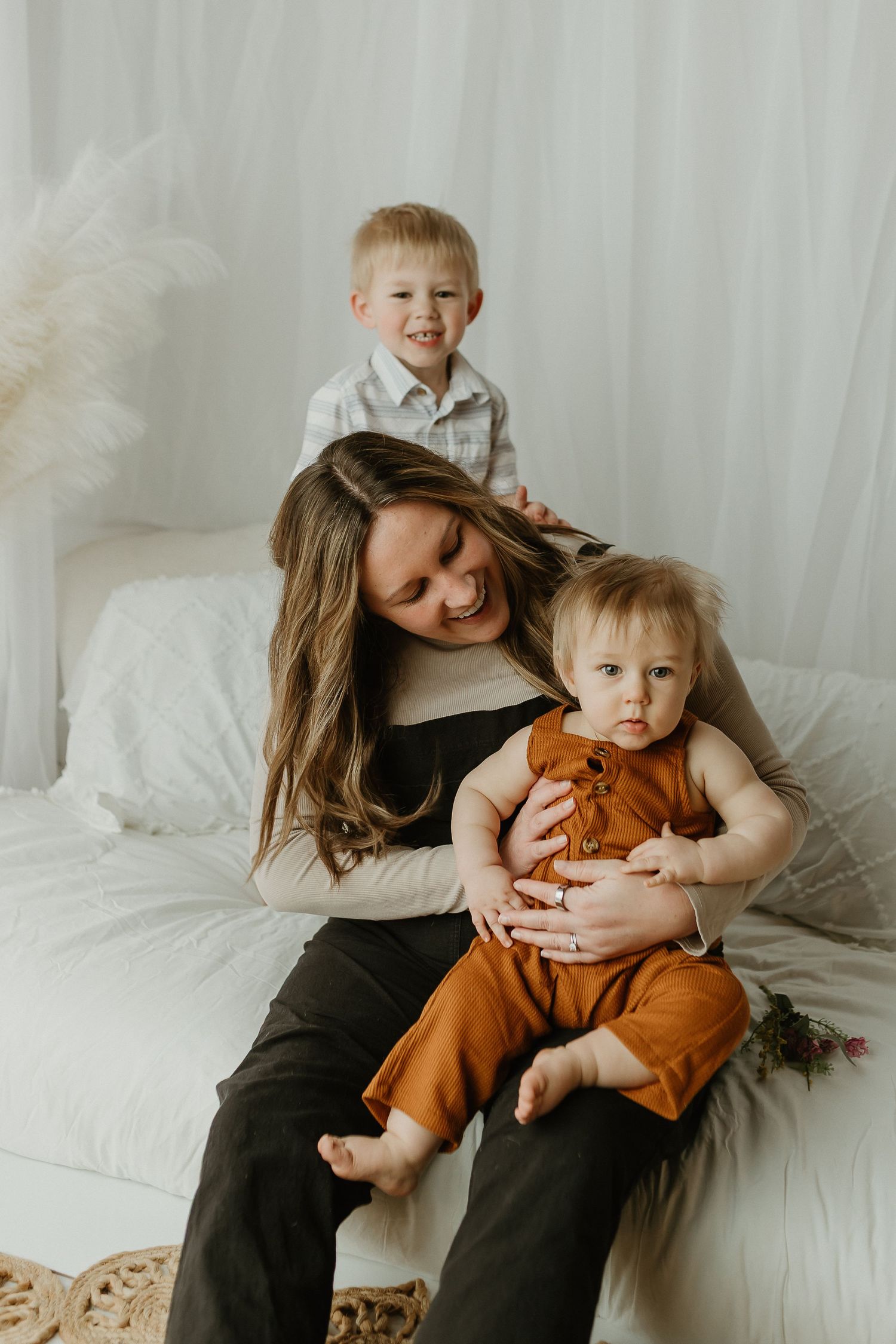 A smiling mother sits on a white bed holding a baby in rust overalls while a toddler boy stands behind her.