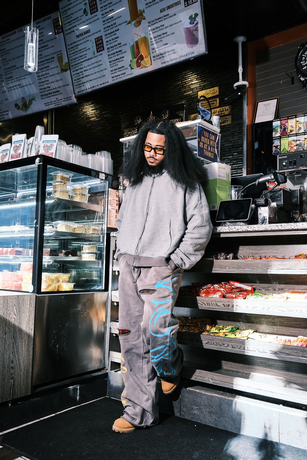Man in gray hoodie and cargo pants stands in a deli shop, wearing glasses and brown boots.