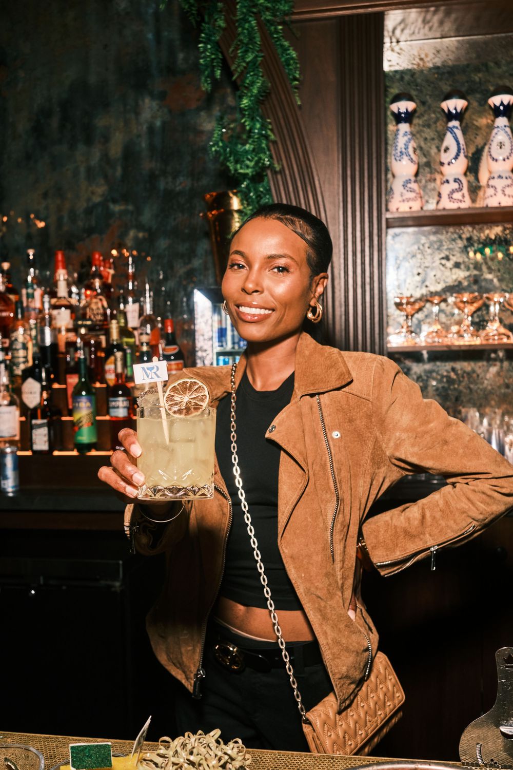 A smiling woman in a brown suede jacket holds a cocktail at a stylish bar with bottles in the background.