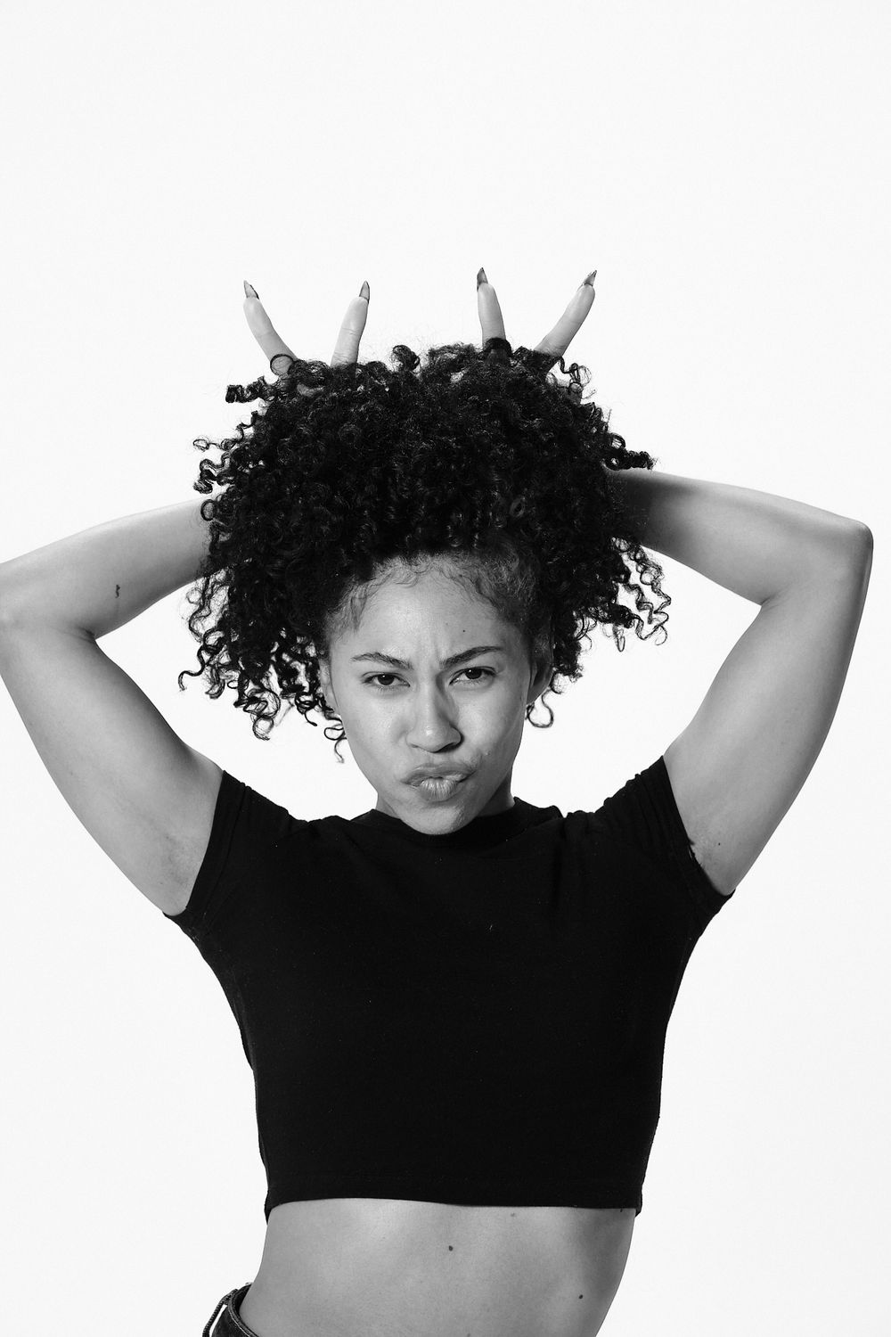 Black and white portrait of a young woman with curly hair making a funny face, hands raised above her head.