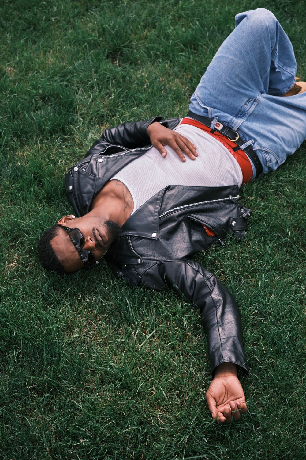 A stylish man in a leather jacket, jeans, and sunglasses relaxing on green grass outdoors.