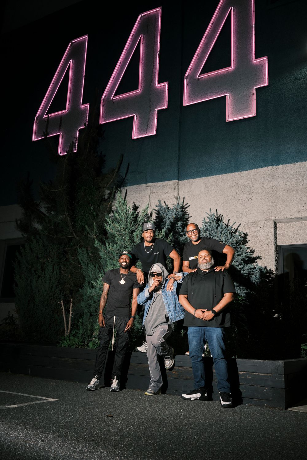 Five people pose at night in front of a building with large pink neon 444 sign and trees.