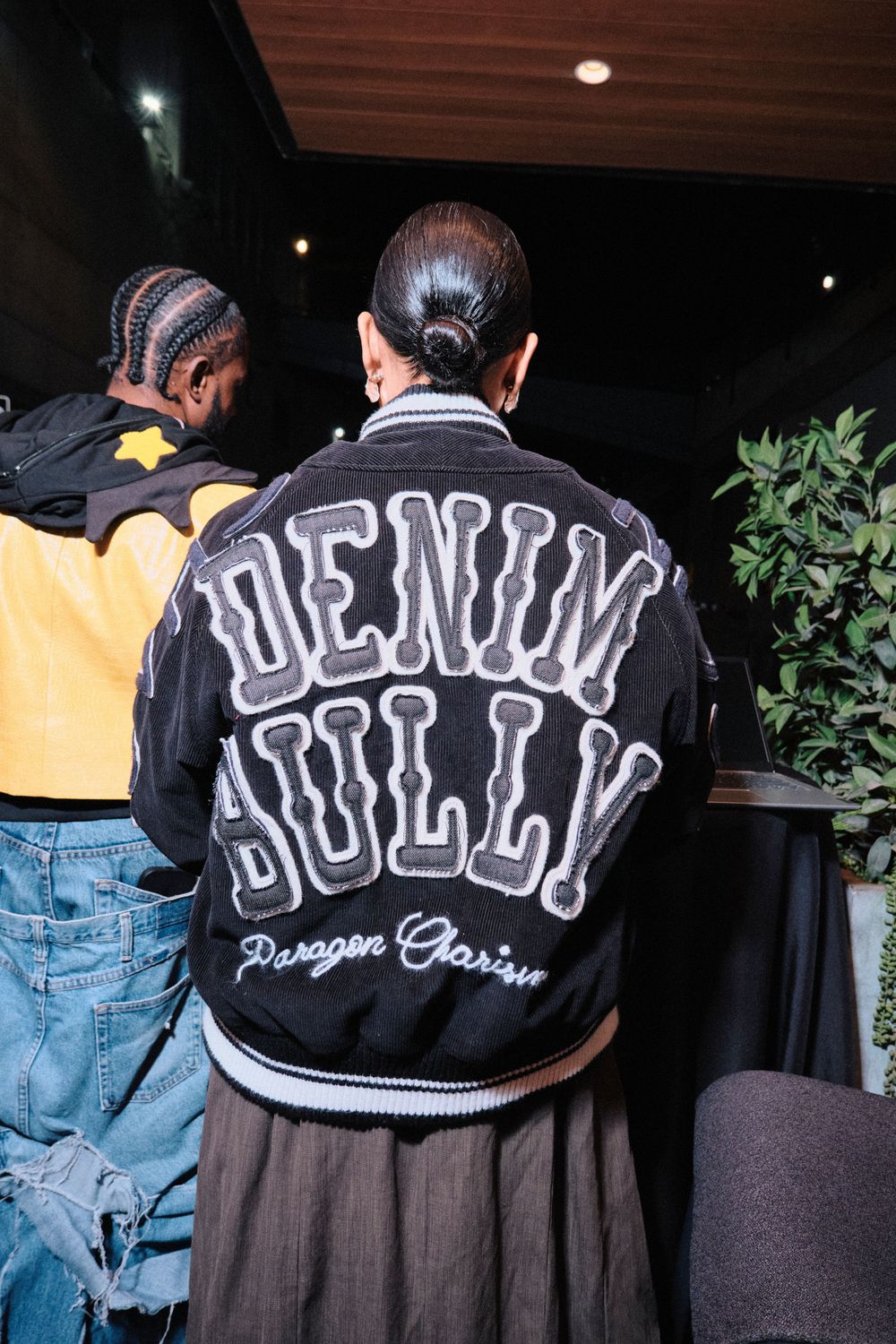 Person wearing a black Denim Bully Paragon Garisu varsity jacket, seen from behind at an event.