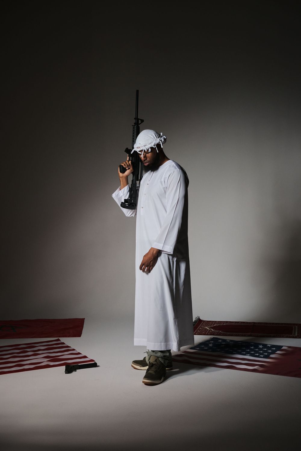 Man in white traditional Middle Eastern attire holding a rifle while standing over American flags on a studio floor.