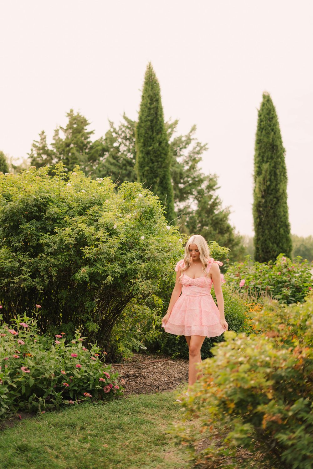 Blonde woman in pink dress walking through lush garden path surrounded by green shrubs and cypress trees.