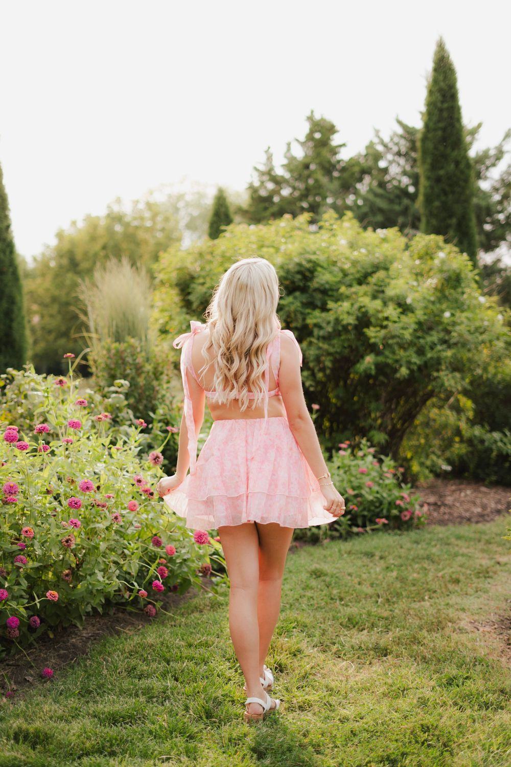 Blonde woman in pink crop top and skirt walking through a lush garden with colorful flowers and green trees.