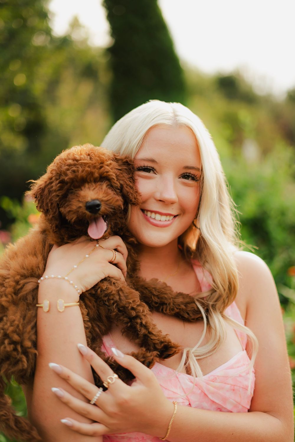 Smiling blonde woman in pink dress hugging an adorable fluffy brown puppy outdoors in a lush green garden.