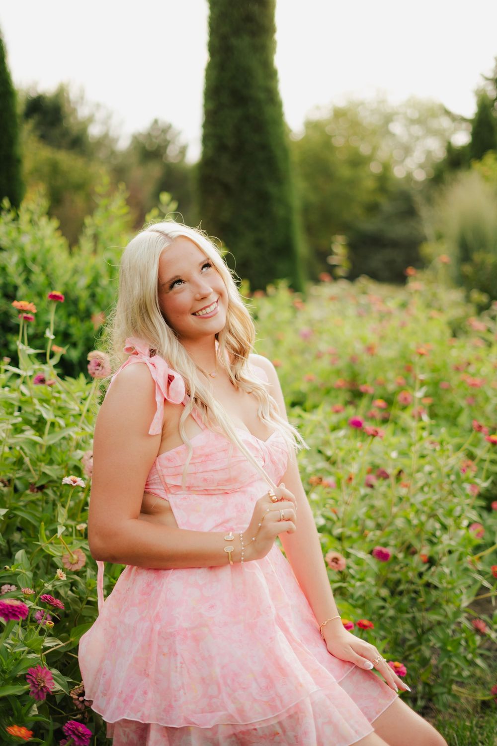 Smiling blonde woman in floral pink dress sitting among colorful zinnia flowers in a garden.
