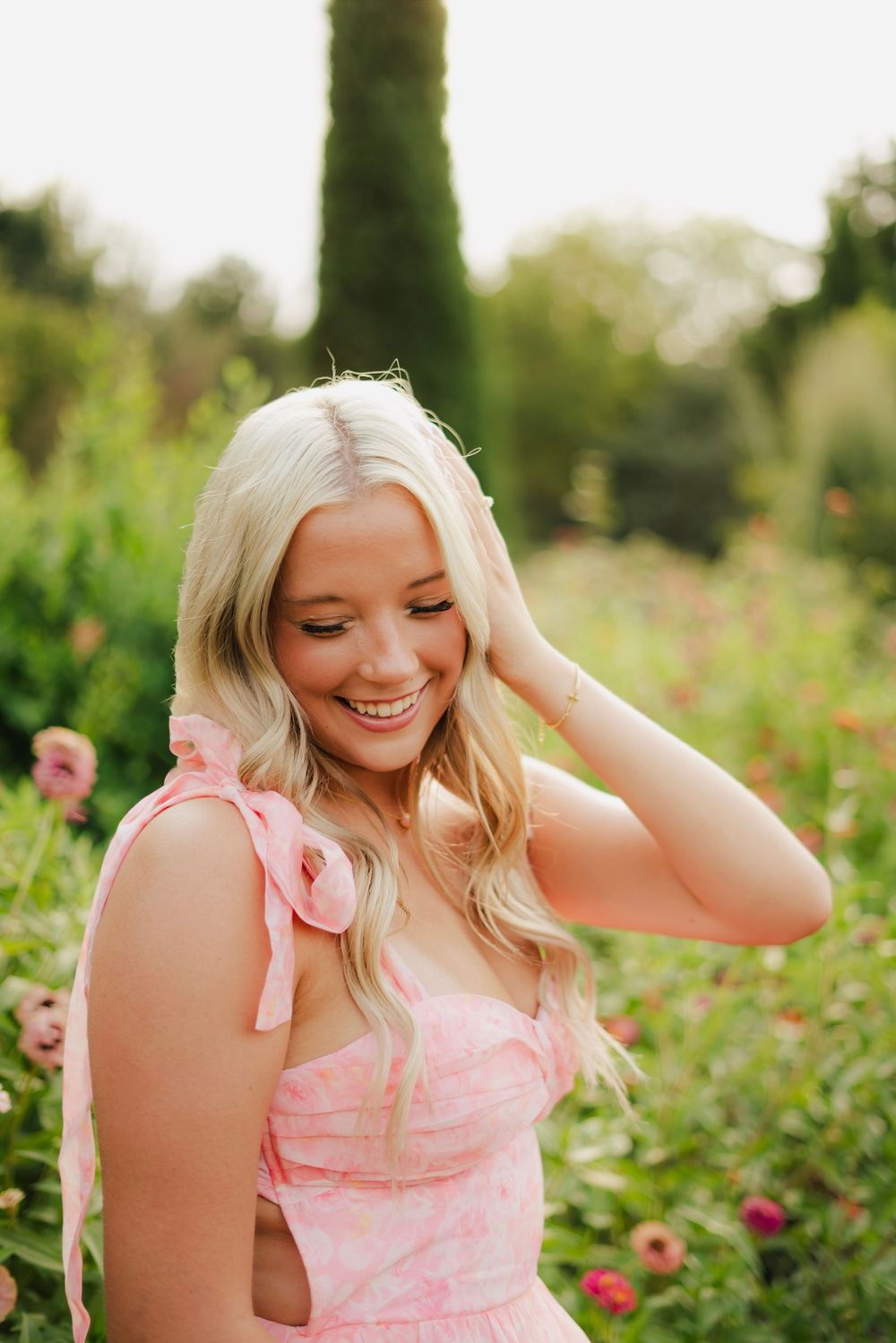 Happy blonde woman in a pink floral sundress laughing in a vibrant flower garden on a sunny day.