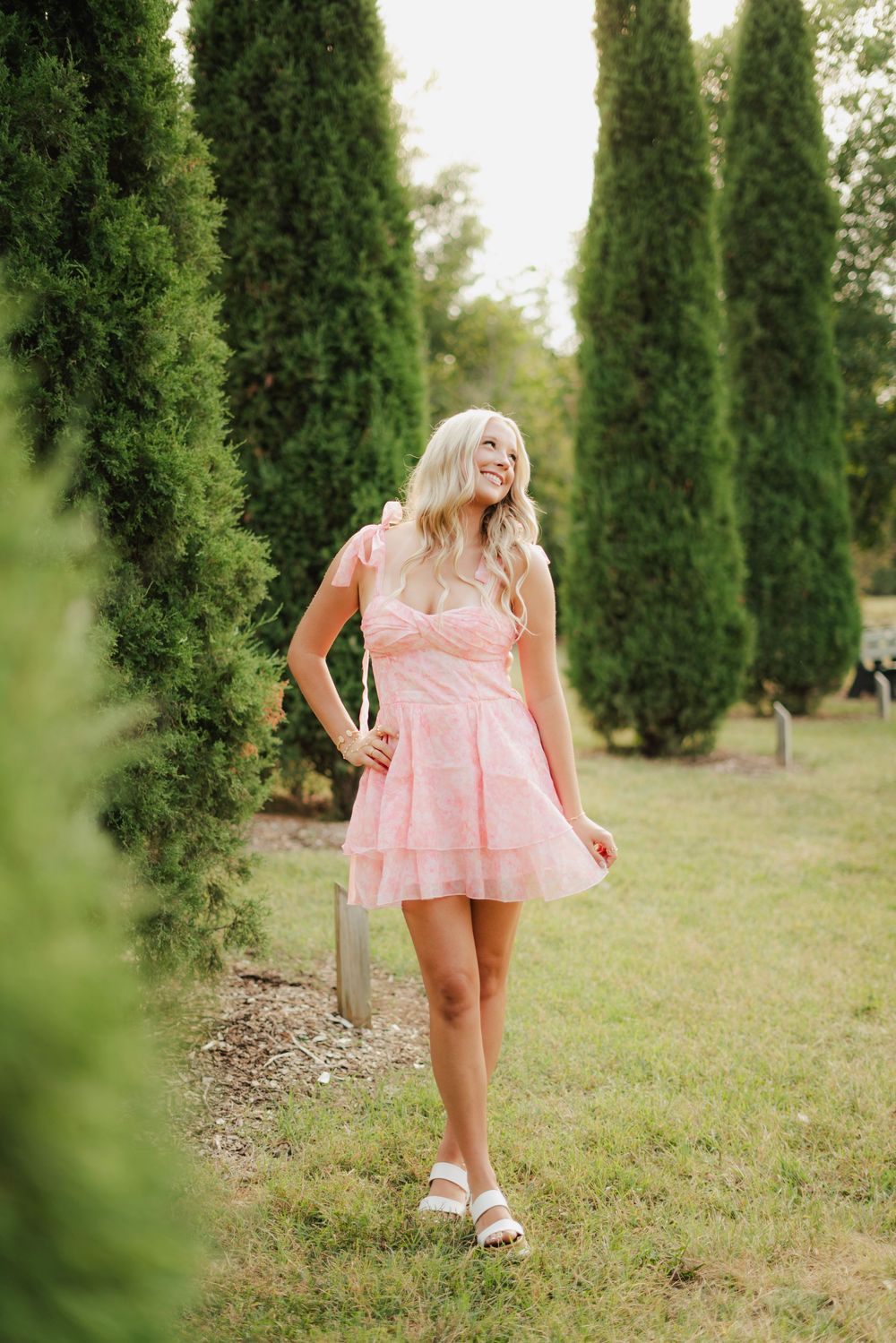 Smiling blonde woman in pink floral mini dress walking among tall cypress trees in a lush green garden.