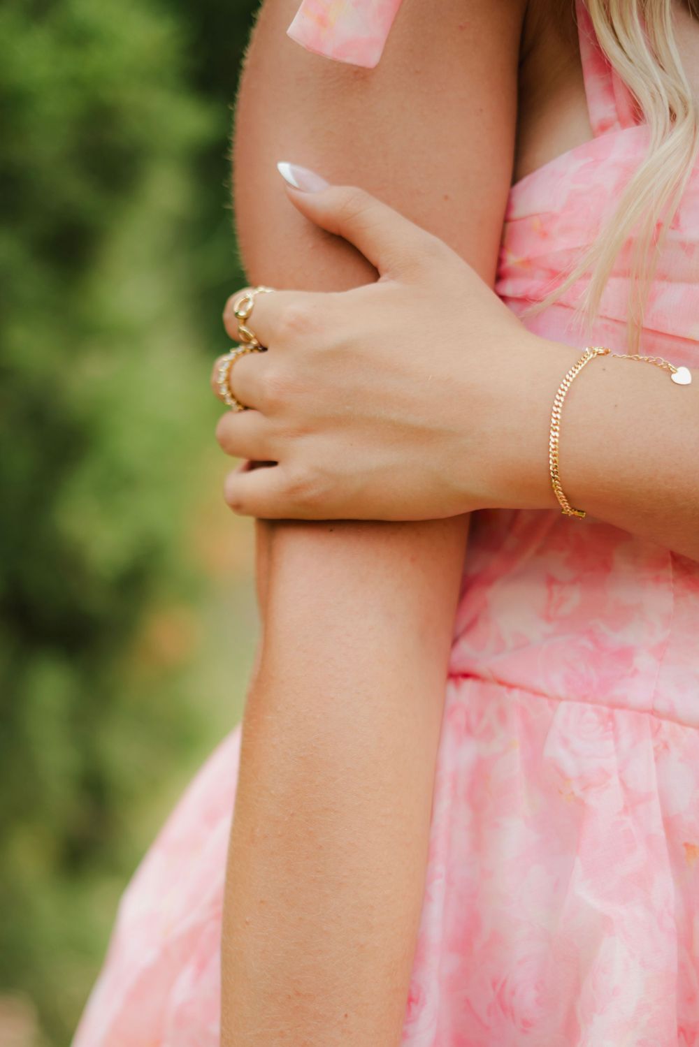 Woman in pink floral dress crossing arms, wearing gold rings and bracelet with white manicured nails.
