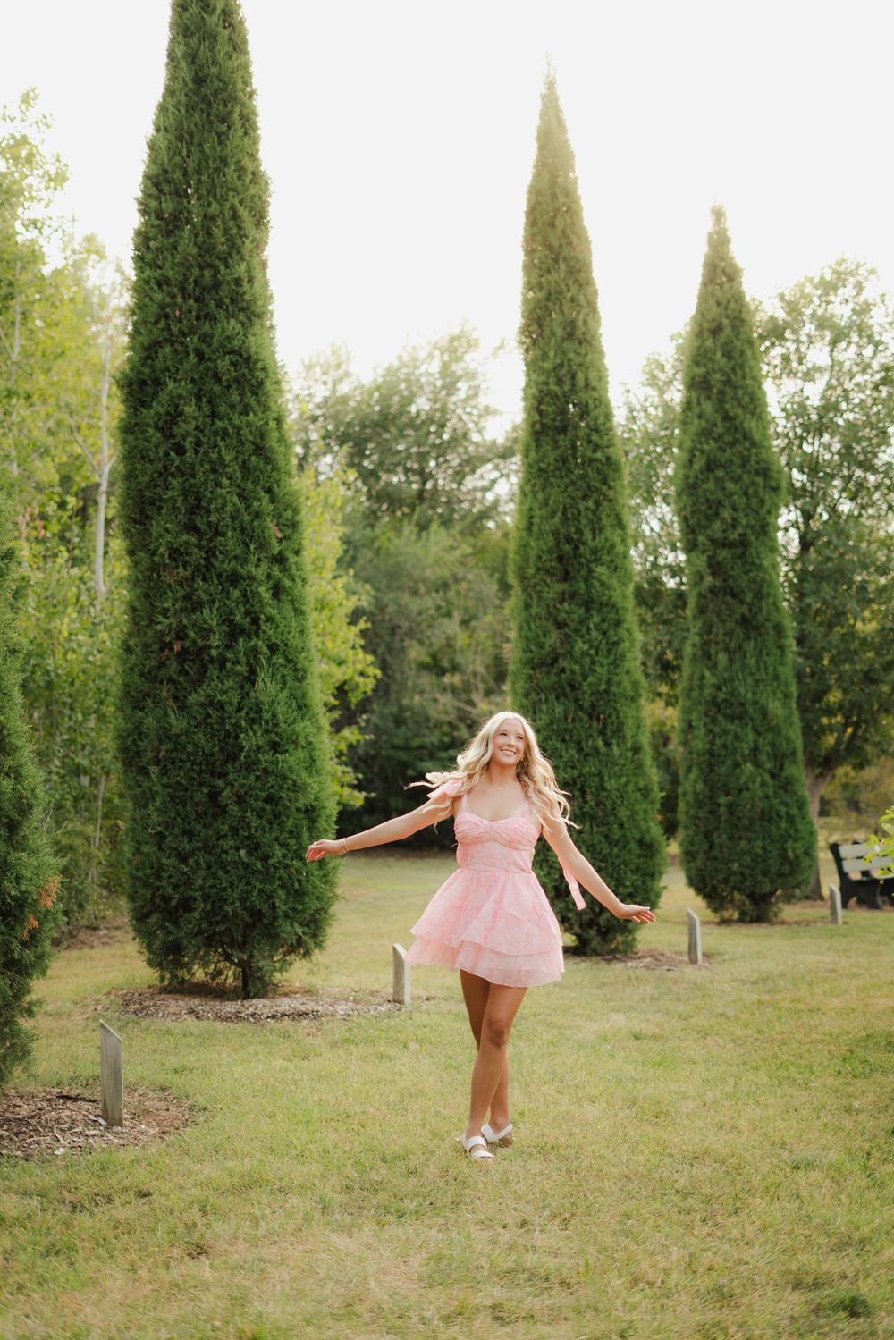 Joyful blonde woman twirling in a pink mini dress between tall cypress trees in a sunny garden.